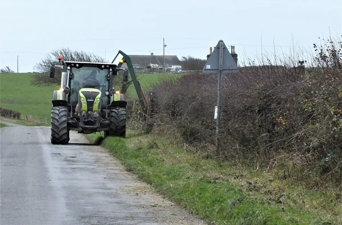 Tractor-mounted hedge cutter trimming a roadside hedge on a country lane